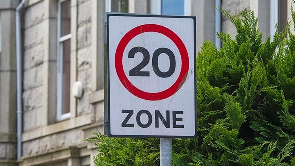 A speed limit sign reads "20 Zone" with a red circle around "20". It's set against lush green bushes and the exterior of an urban stone building.