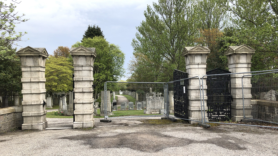 Stone pillars and a closed metal gate block the entrance to a cemetery with lush green trees and tombstones visible in the background, conveying a serene yet solemn atmosphere.