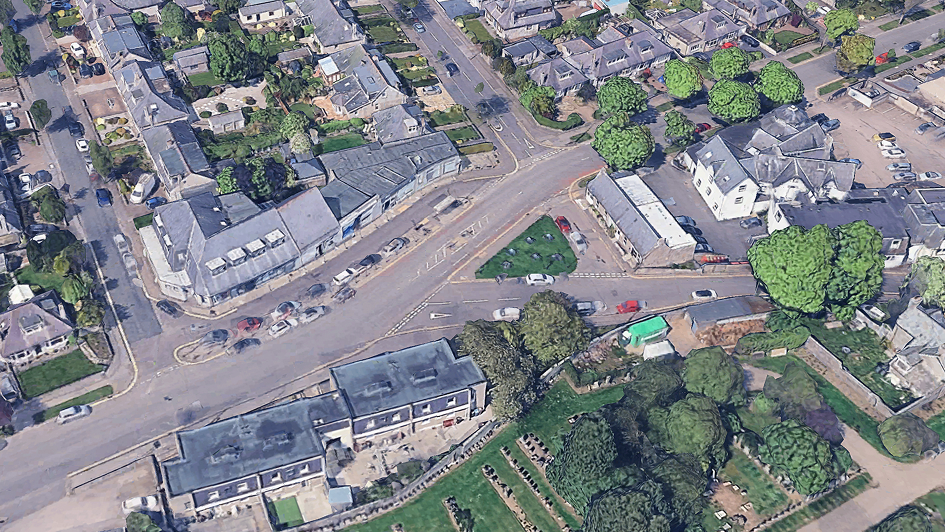 Aerial view of a suburban intersection with houses, parked cars, trees, and green spaces on a clear day.