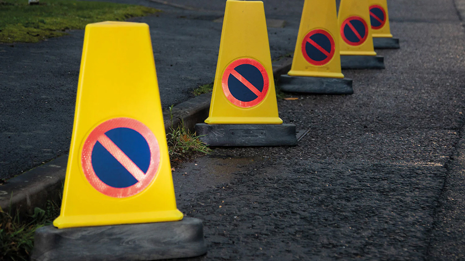 A row of yellow traffic cones with no-parking signs is lined along a wet road with grass edging. The scene conveys caution and restriction.
