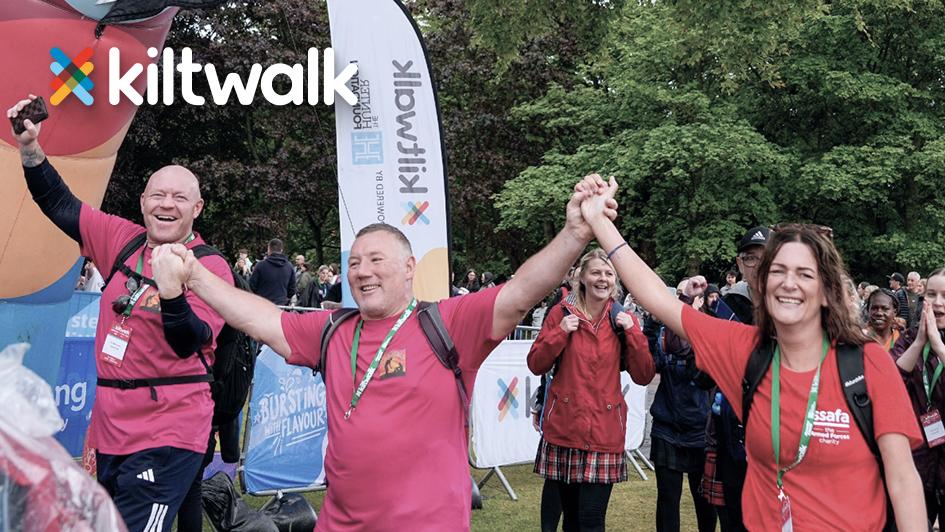 Participants in matching pink shirts celebrate while holding hands at a Kiltwalk event. Banners and a cheering crowd are visible in the background.