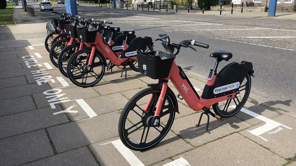 Row of bright red rental bikes parked on a city sidewalk labeled "CYCLE HIRE ONLY." Sunlit pavement and street in the background, conveying urban mobility.