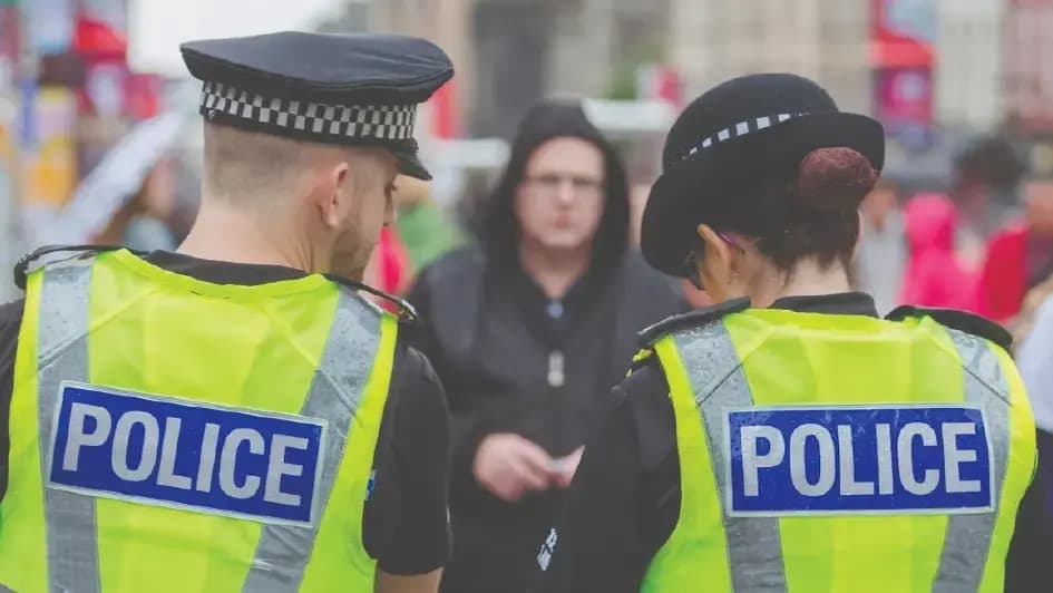 Two police officers in yellow vests, one male, one female, stand in an urban setting. The focus is on their uniforms, with a blurred crowd behind.