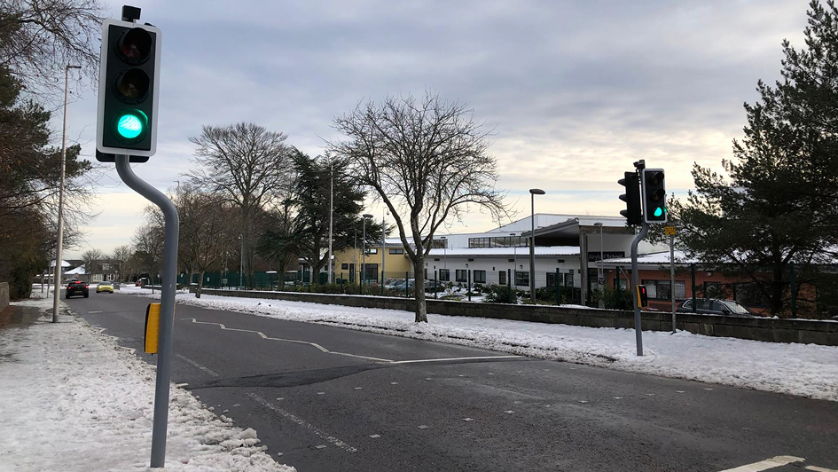 A snow-lined road with two active green traffic lights under a cloudy sky. Sparse traffic and bare trees create a calm winter atmosphere.